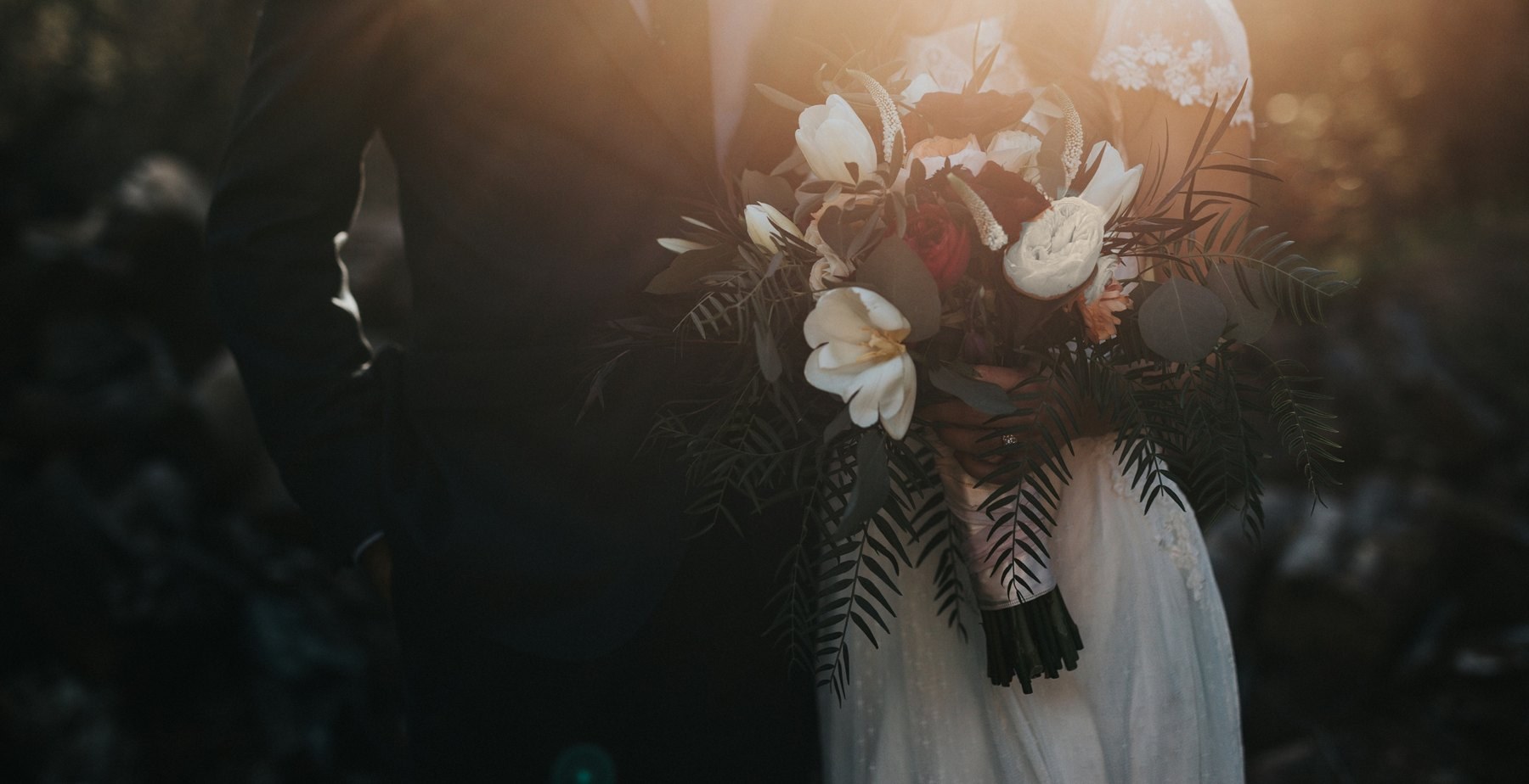 A bride holding a bouquet in warm evening light, beside a guest in a dark suit