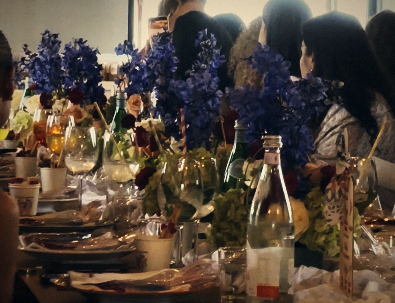 A long celebration table with blue and red flowers, glasses and guests seated together
